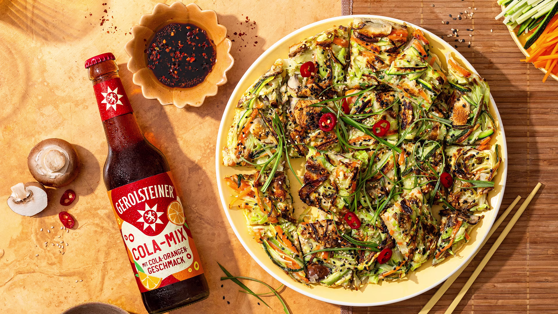 Draufsicht auf koreanische Gemüsepfannkuchen mit Frühlingszwiebeln, Sesam und Chili auf einem Teller neben einer Flasche Gerolsteiner Cola-Mix. Dazu werden Sojasauce-Dip und Essstäbchen serviert.