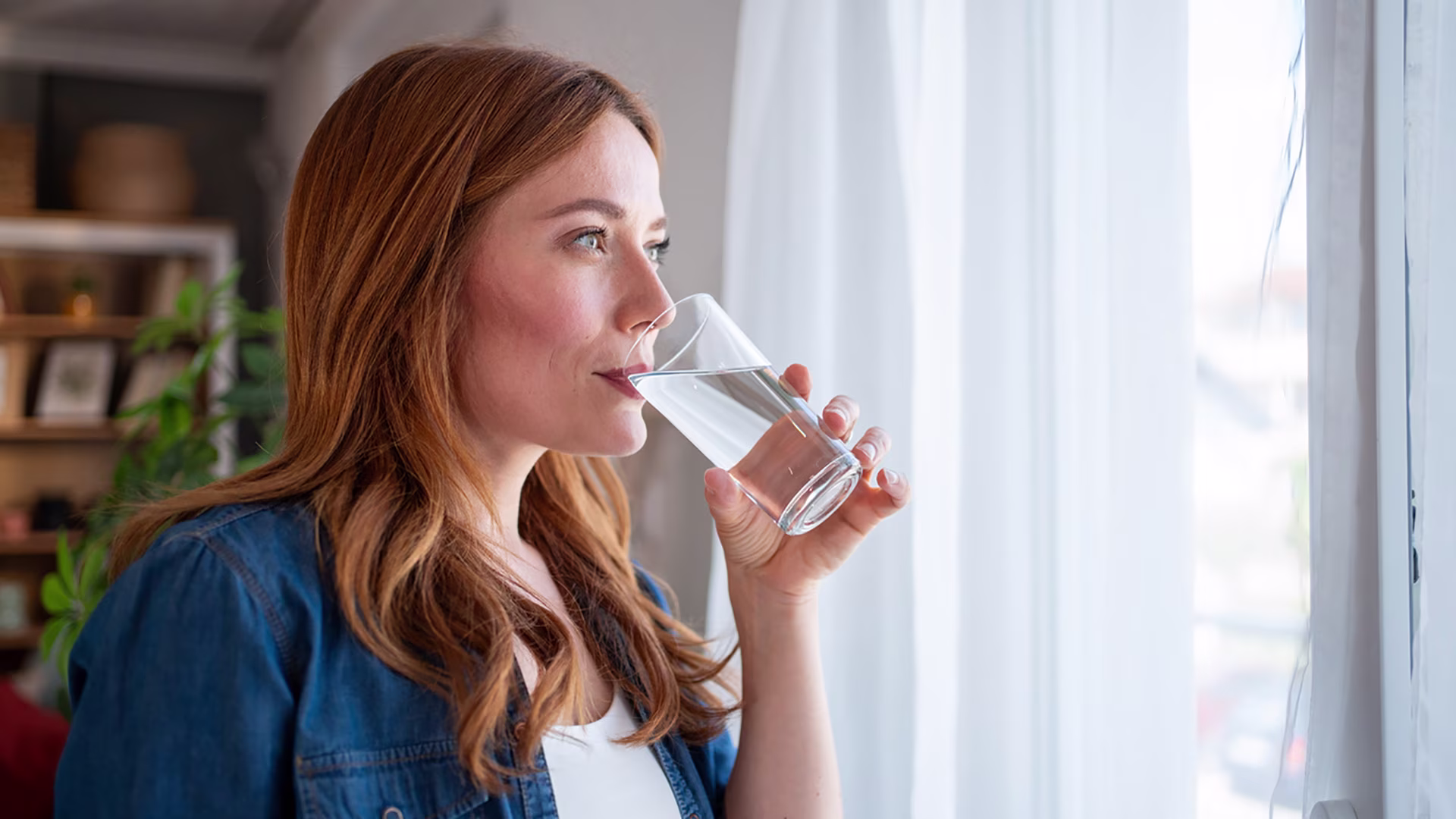 Frau trinkt ein Glas Wasser am Fenster zur Unterstützung der Verdauung bei Blähbauch.