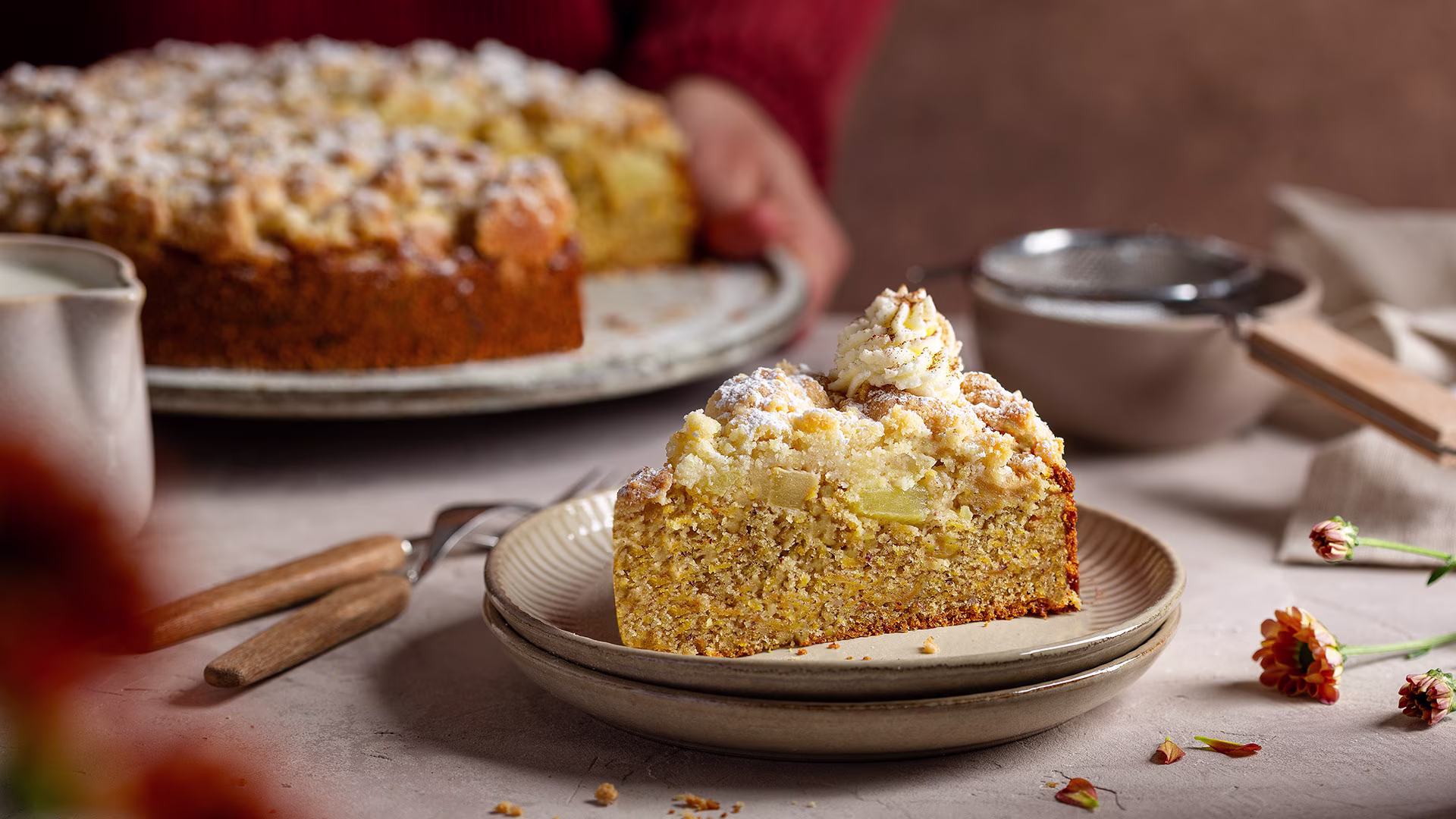 Ein Stück Kürbis-Apfel-Streuselkuchen mit Puderzucker und einem Tupfer Sahne auf einem Teller. Im Hintergrund wird der Kuchen serviert, daneben stehen ein Milchkännchen und ein Sieb mit Puderzucker – herbstliche Kaffeetafel.