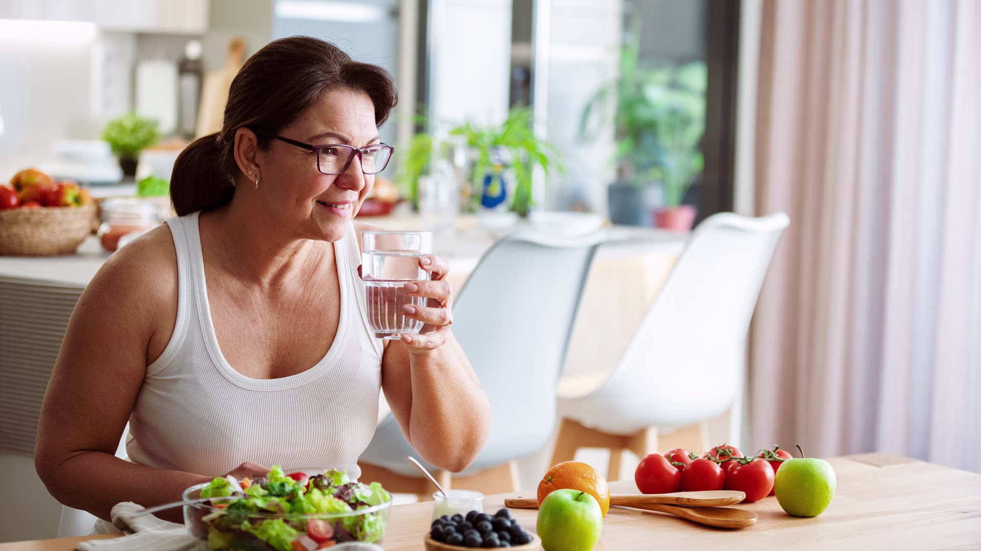 Une femme boit un verre d'eau en mangeant une salade et des fruits.
