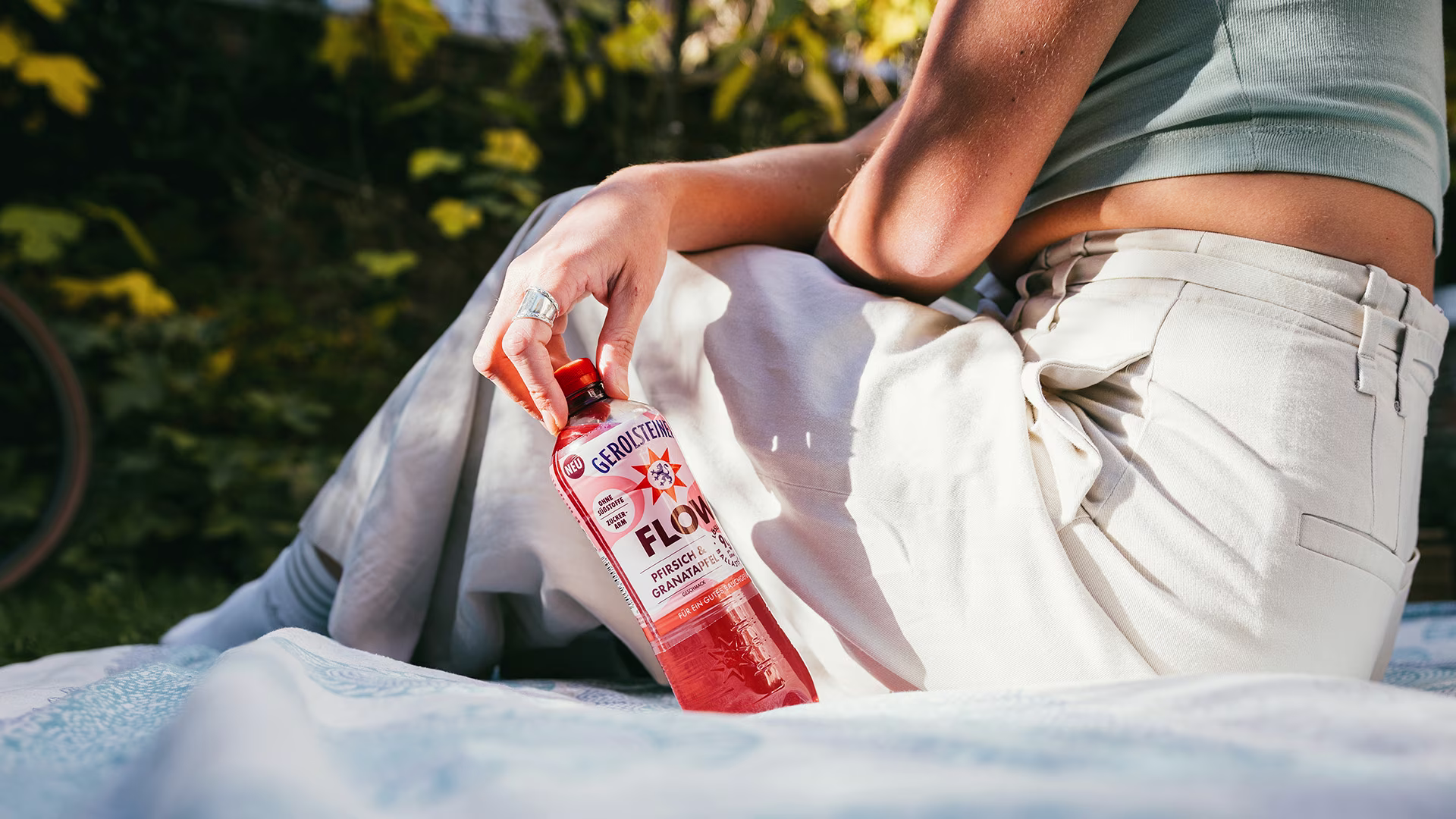 Eine Person sitzt entspannt auf einer Decke im Garten und hält eine Flasche Gerolsteiner Flow Pfirsich & Granatapfel in der Hand. Sonnenlicht und grüne Pflanzen erzeugen eine sommerlich-entspannte Stimmung.