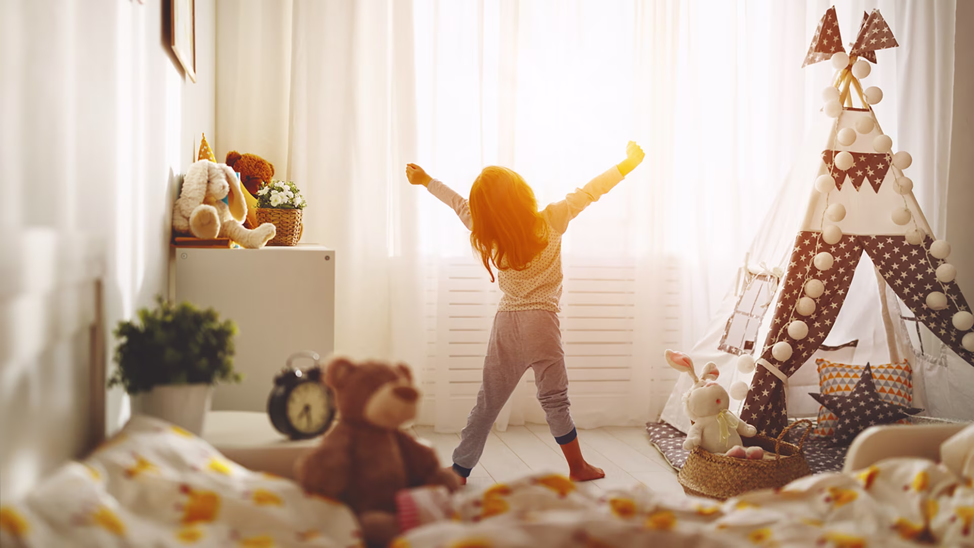 Kleines Mädchen streckt sich morgens nach dem Aufstehen. Die Sonne scheint ins Zimmer.