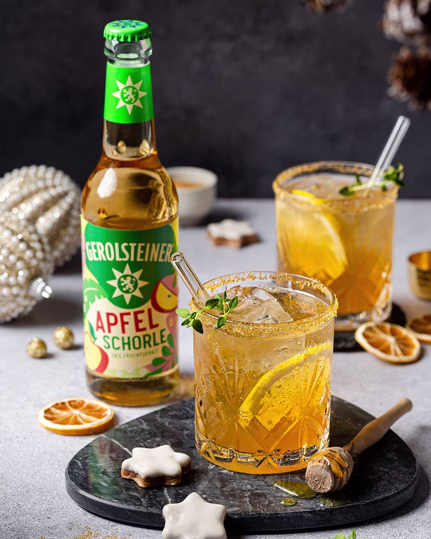 Eine Flasche Gerolsteiner Apfelschorle steht neben zwei Gläsern mit goldgelbem Getränk, Eis und Orangenscheiben. Dekoration mit Honiglöffel, Zimtsternen und goldenen Kugeln sorgt für eine festliche, winterliche Atmosphäre.