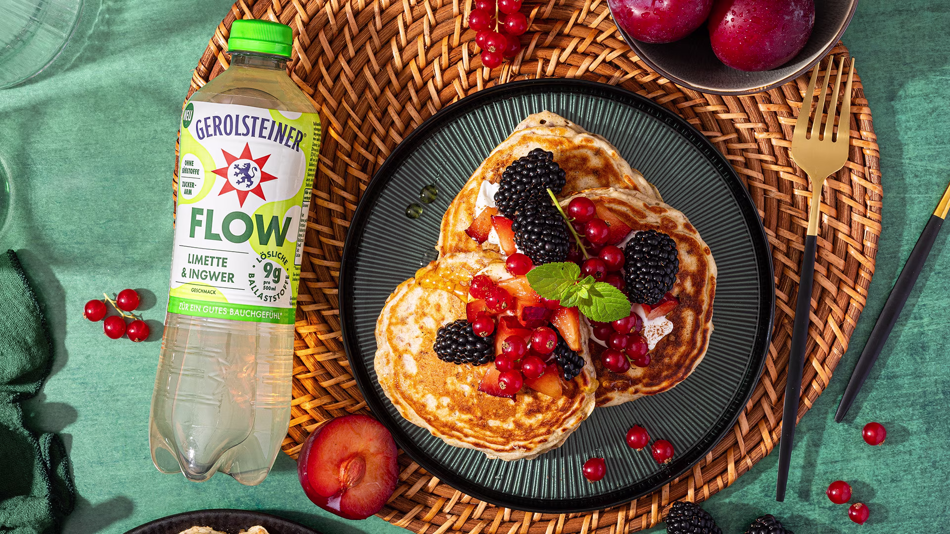 Müslipancakes mit Brombeeren, Johannisbeeren, Pflaumen und Minze auf einem dunklen Teller. Daneben steht eine Flasche Gerolsteiner Flow Limette & Ingwer.