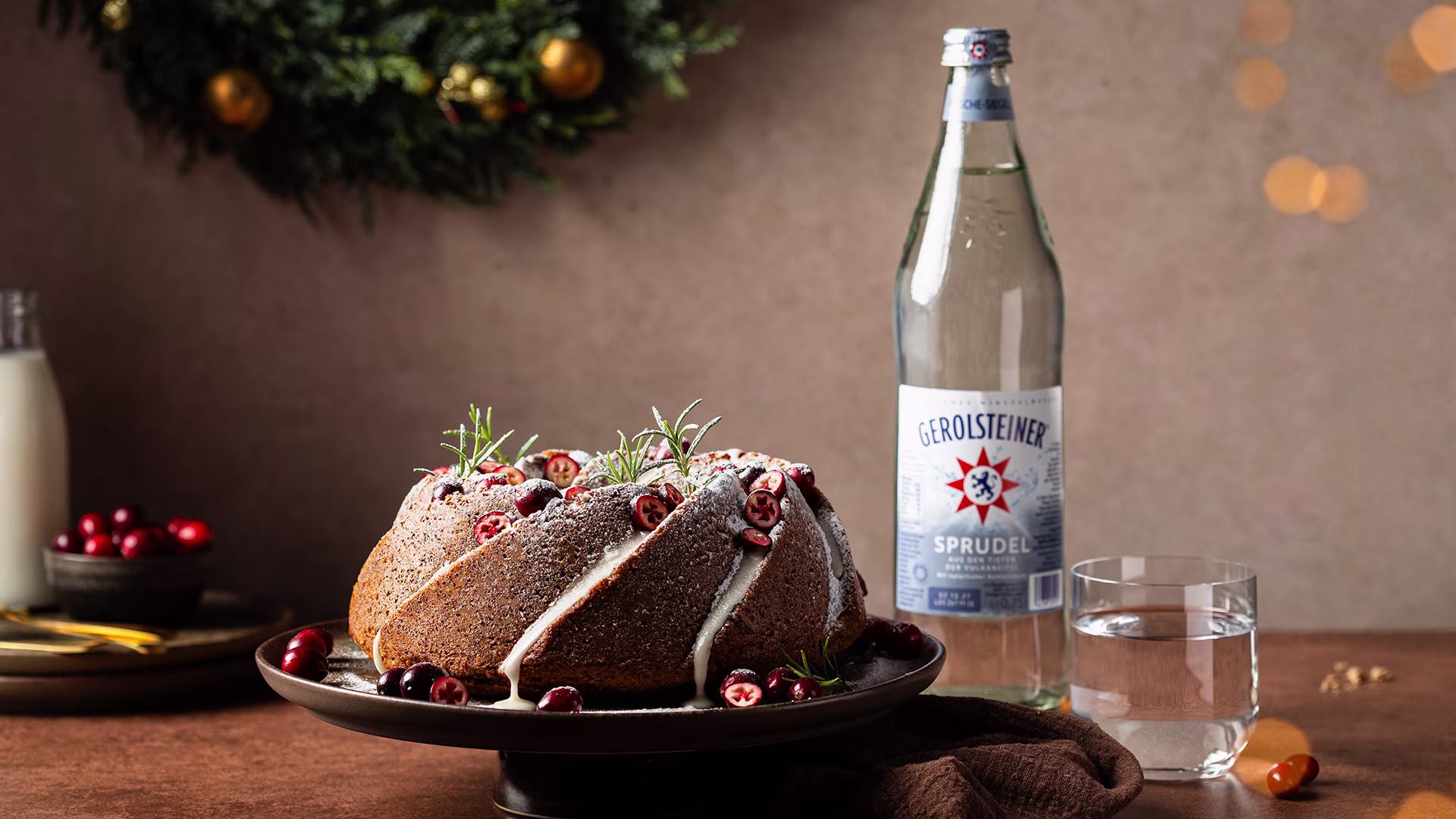 Ein winterlicher Nuss-Gugelhupf mit Zuckerguss, Cranberries und Rosmarin steht auf einem Teller. Daneben eine Flasche Gerolsteiner Sprudel und ein Glas Wasser. Im Hintergrund sorgen Lichter und ein Adventskranz für festliche Stimmung.