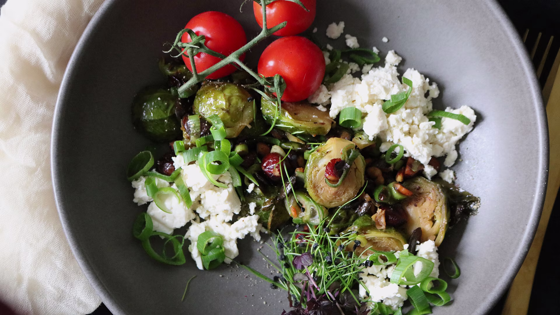 Ein Teller mit gebratenem Rosenkohl, Kirschtomaten, Feta, Frühlingszwiebeln und frischen Kräutern. Das bunte Gericht ist ansprechend angerichtet und vermittelt eine frische, gesunde und mediterrane Atmosphäre.