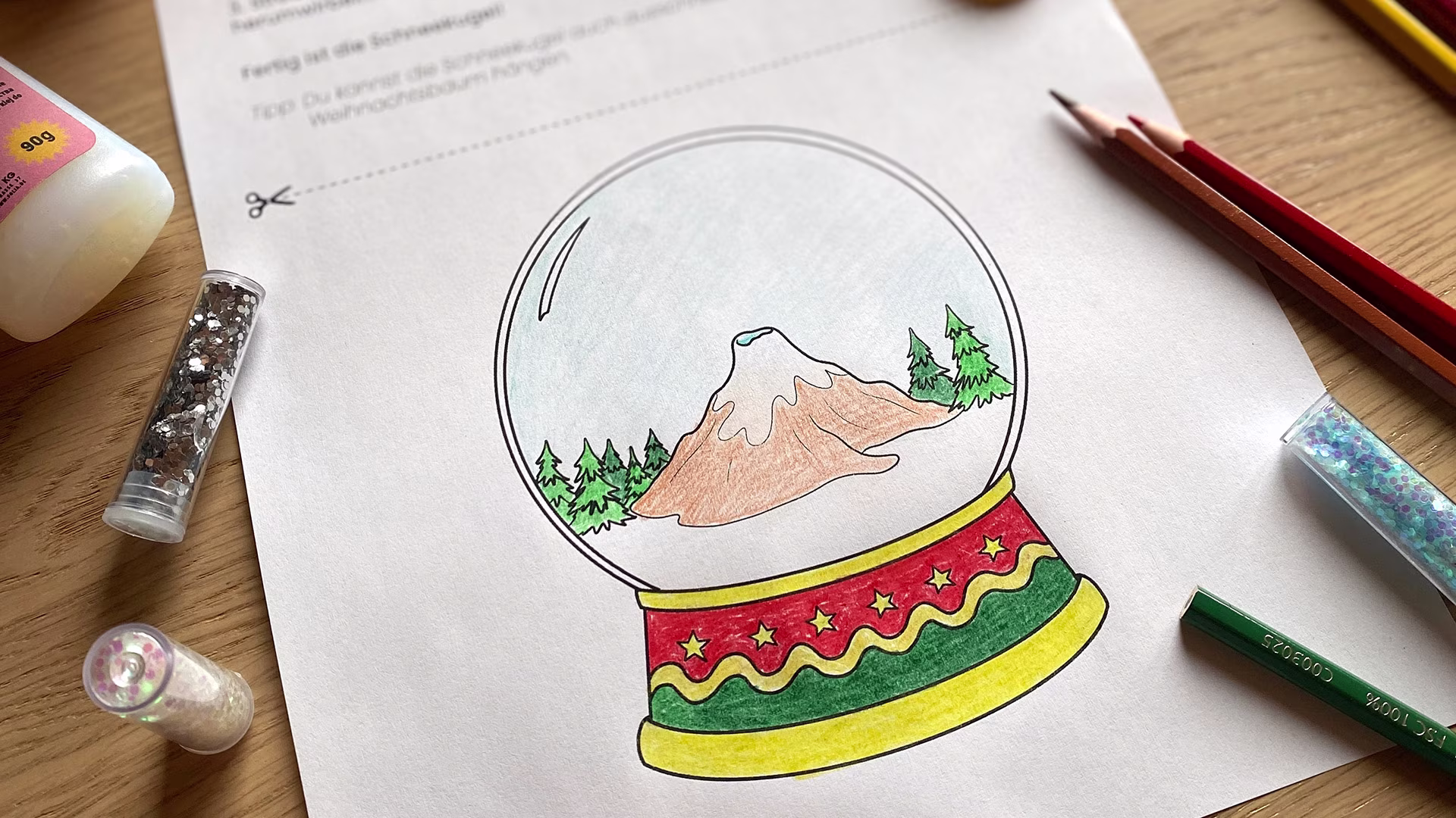 Eine ausgemalte Schneekugel mit Berglandschaft und Tannen steht im Zentrum eines Bastelbogens. Drumherum liegen Buntstifte, Glitzer und Bastelmaterial. Die Szene wirkt kreativ und weihnachtlich.