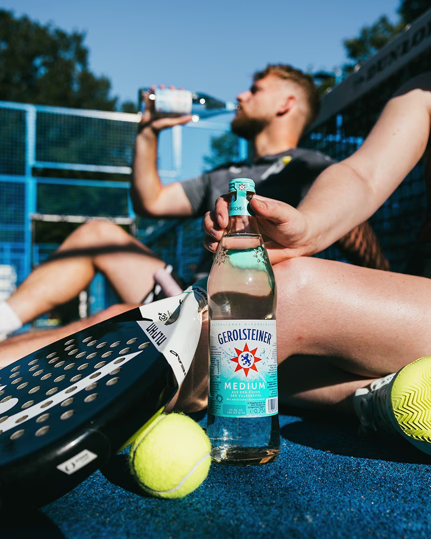 Gerolsteiner Medium Mineralwasser auf einem Padelplatz zwischen Schlägern und Tennisbällen. Im Hintergrund trinken zwei Personen nach dem Spiel Wasser und entspannen auf dem Platz bei sonnigem Wetter.