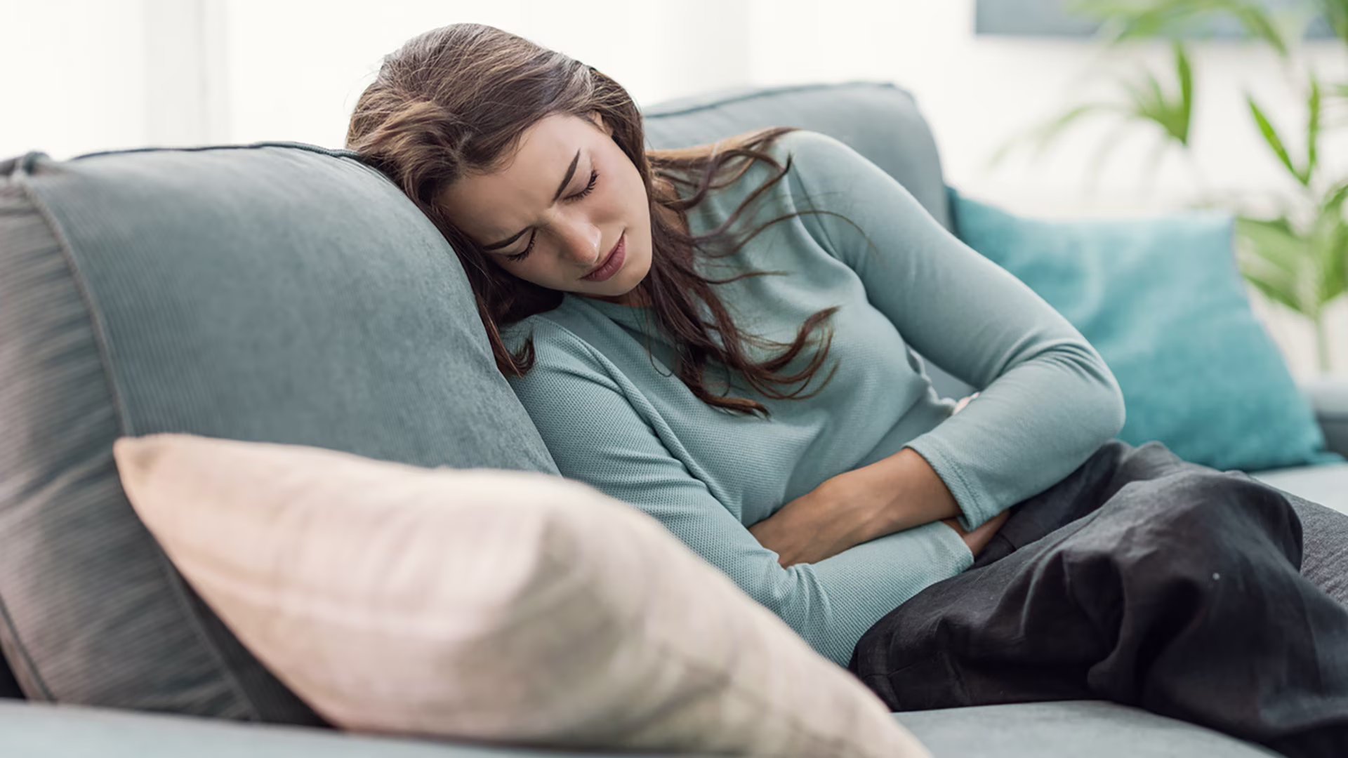 : Junge Frau liegt mit schmerzverzerrtem Gesicht auf dem Sofa und hält sich den Bauch, leidend unter Bauchschmerzen oder Blähungen.