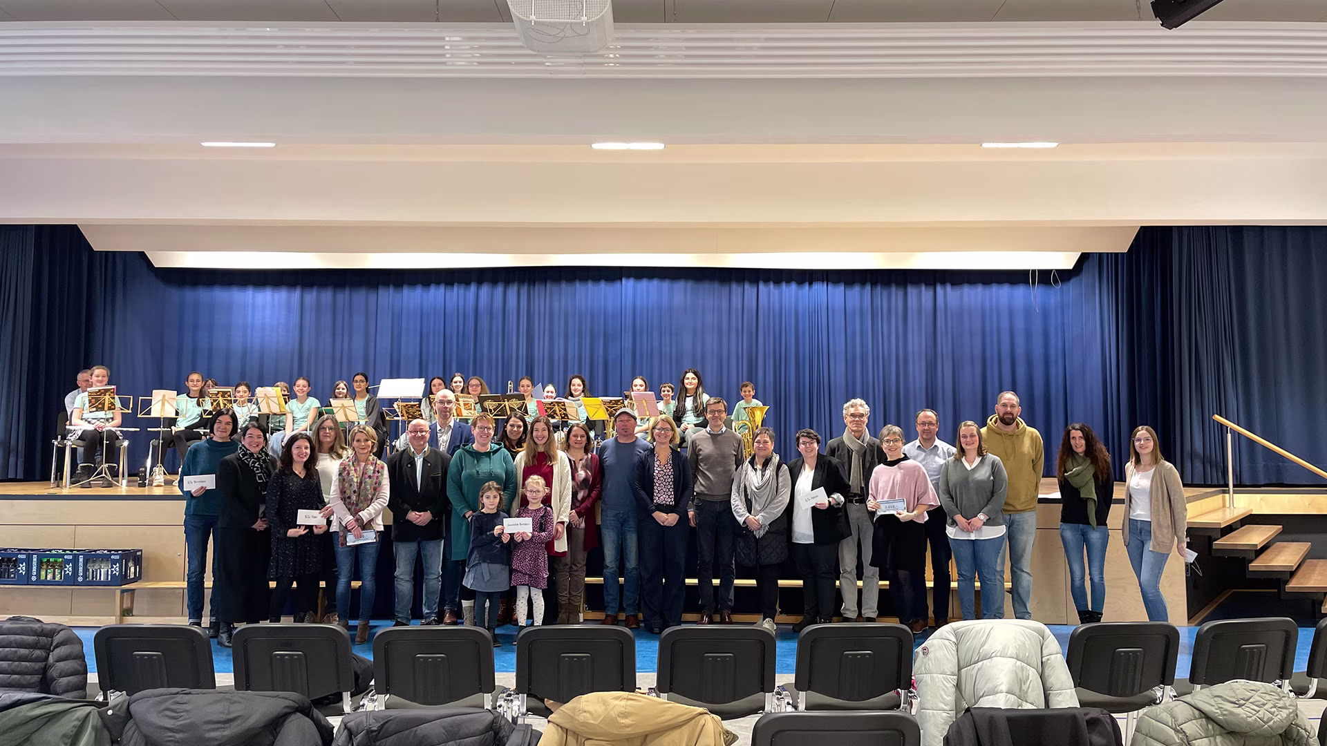Gruppenfoto auf einer Schulaula-Bühne mit Kindern eines Schulorchesters im Hintergrund und Erwachsenen aus verschiedenen Institutionen im Vordergrund bei der feierlichen Übergabe der Gerolsteiner Weihnachtsspende 2025.