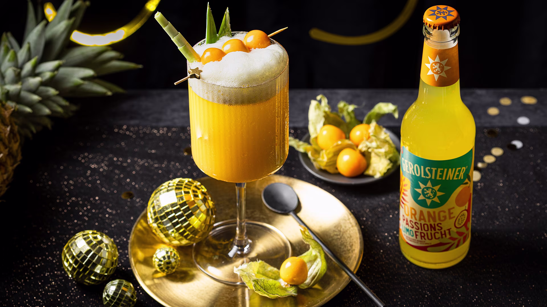 Ein tropischer Mocktail mit Ananasblättern und Physalis steht auf einem goldenen Teller. Daneben eine Flasche Gerolsteiner Orange Passionsfrucht. Goldene Deko-Kugeln und dunkler Hintergrund erzeugen eine festlich-exotische Stimmung.