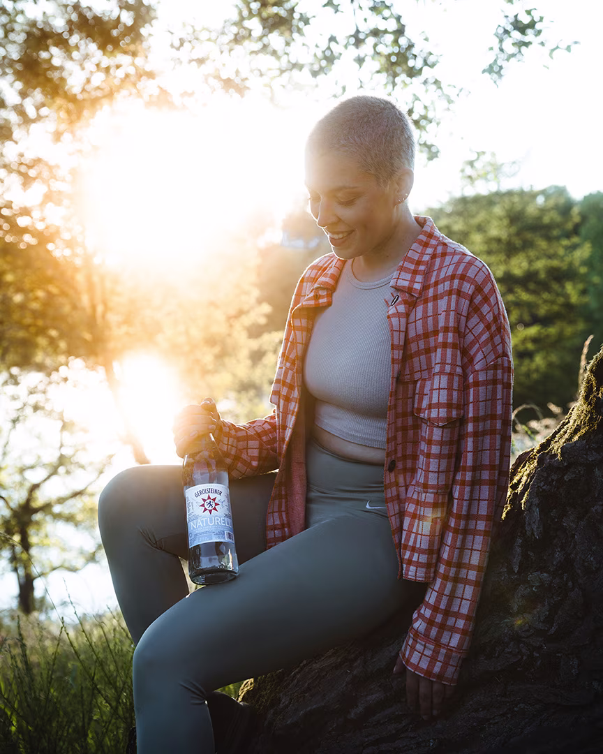Mann in Freizeitkleidung sitzt bei Sonnenuntergang auf einem Baumstamm in der Natur und hält eine Flasche Gerolsteiner Naturell in der Hand.