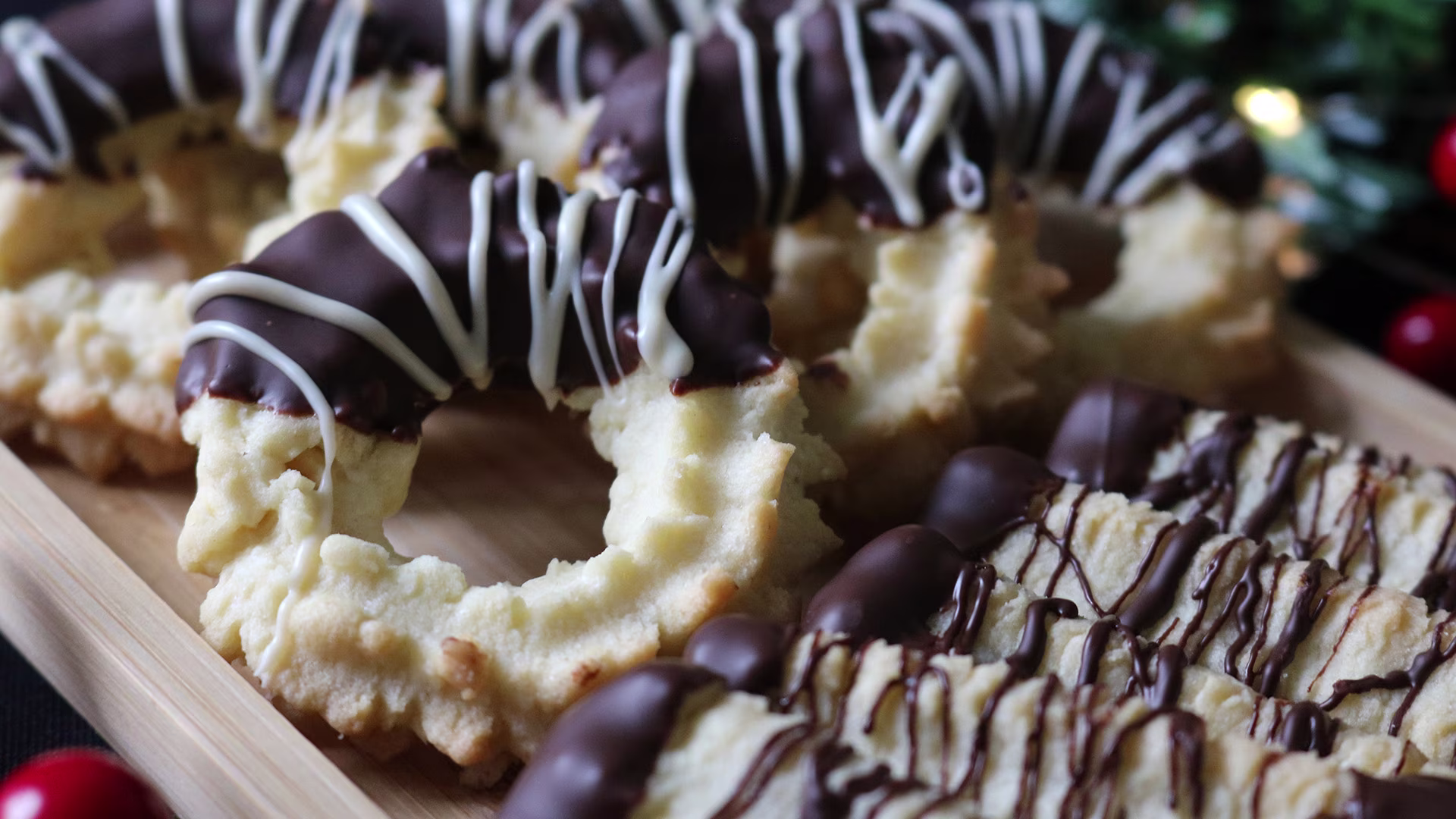 Auf einem Holztablett liegt frisch gebackenes Spritzgebäck, teils mit dunkler und weißer Schokolade überzogen und verziert. Im Hintergrund sorgen weihnachtliche Dekorationen für eine festliche Stimmung.