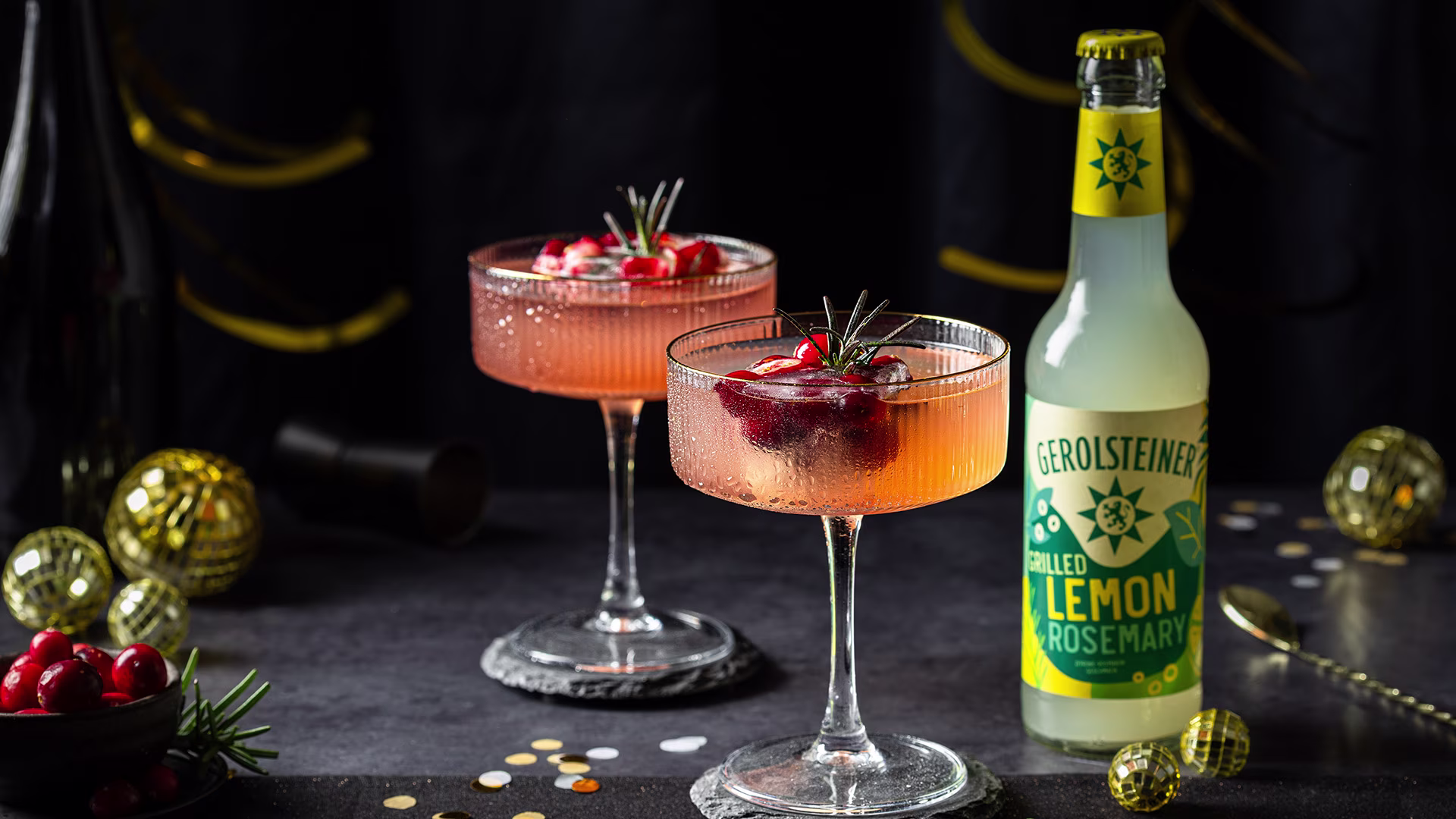 Zwei festliche Cocktails in eleganten Gläsern stehen auf einem dunklen Tisch. Sie sind mit Cranberries und Rosmarin garniert. Daneben eine Flasche Gerolsteiner Grilled Lemon Rosemary. Goldene Deko sorgt für eine stimmungsvolle Atmosphäre.