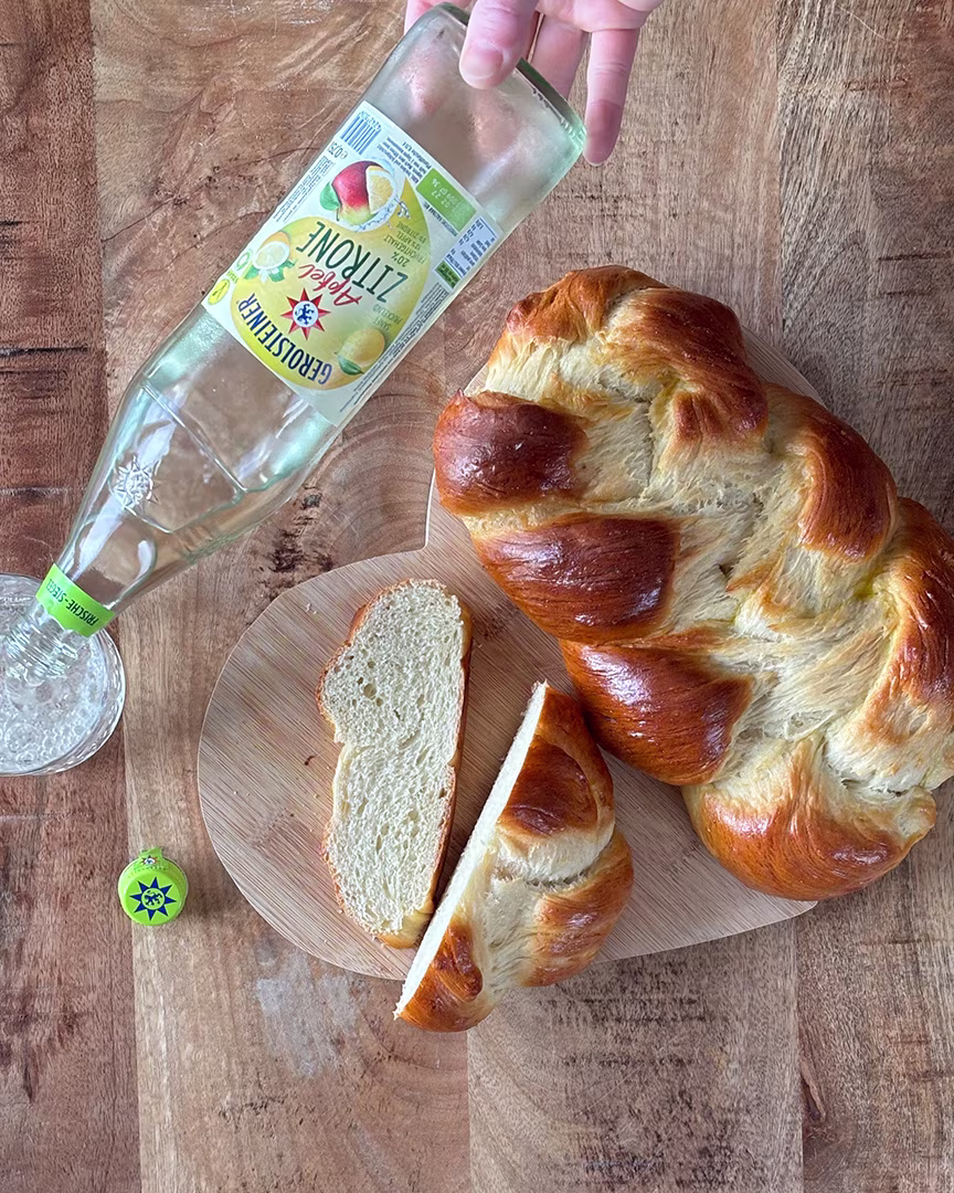 Ein frisch gebackener Hefezopf liegt auf einem Holzbrett, daneben eine aufgeschnittene Scheibe. Dazu wird ein Glas Gerolsteiner Apfel-Zitrone eingeschenkt – eine fruchtig-frische Begleitung zum süßen Gebäck.