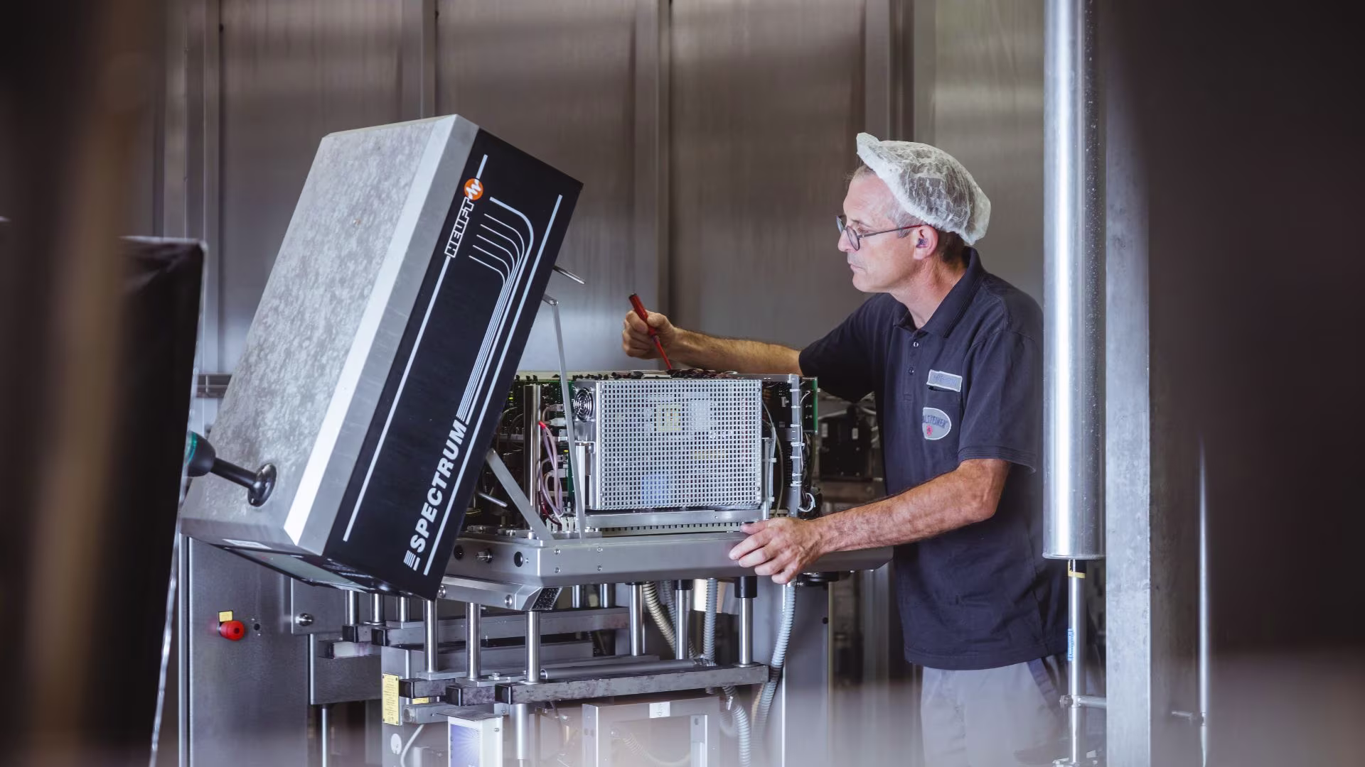 Zwei Mitarbeiter bei der technischen Wartung einer Industrieanlage; einer arbeitet konzentriert mit Werkzeug, der andere reicht ein Messinstrument – beide in Arbeitskleidung mit Haarnetz bzw. Warnweste.