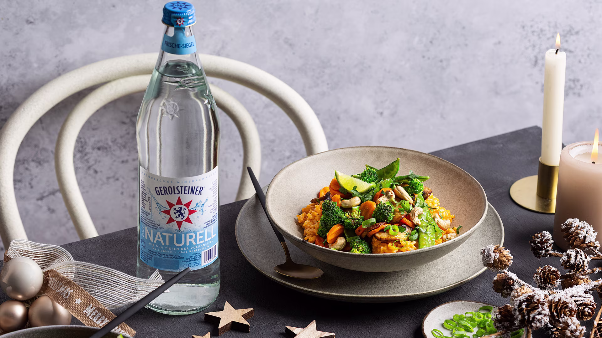 Schale mit cremigem Thai-Curry-Risotto, garniert mit Brokkoli, Zuckerschoten, Karotten, Cashewkernen und Limette. Daneben steht eine Flasche Gerolsteiner Naturell. Kerzen und winterliche Dekoration schaffen eine gemütliche Atmosphäre.