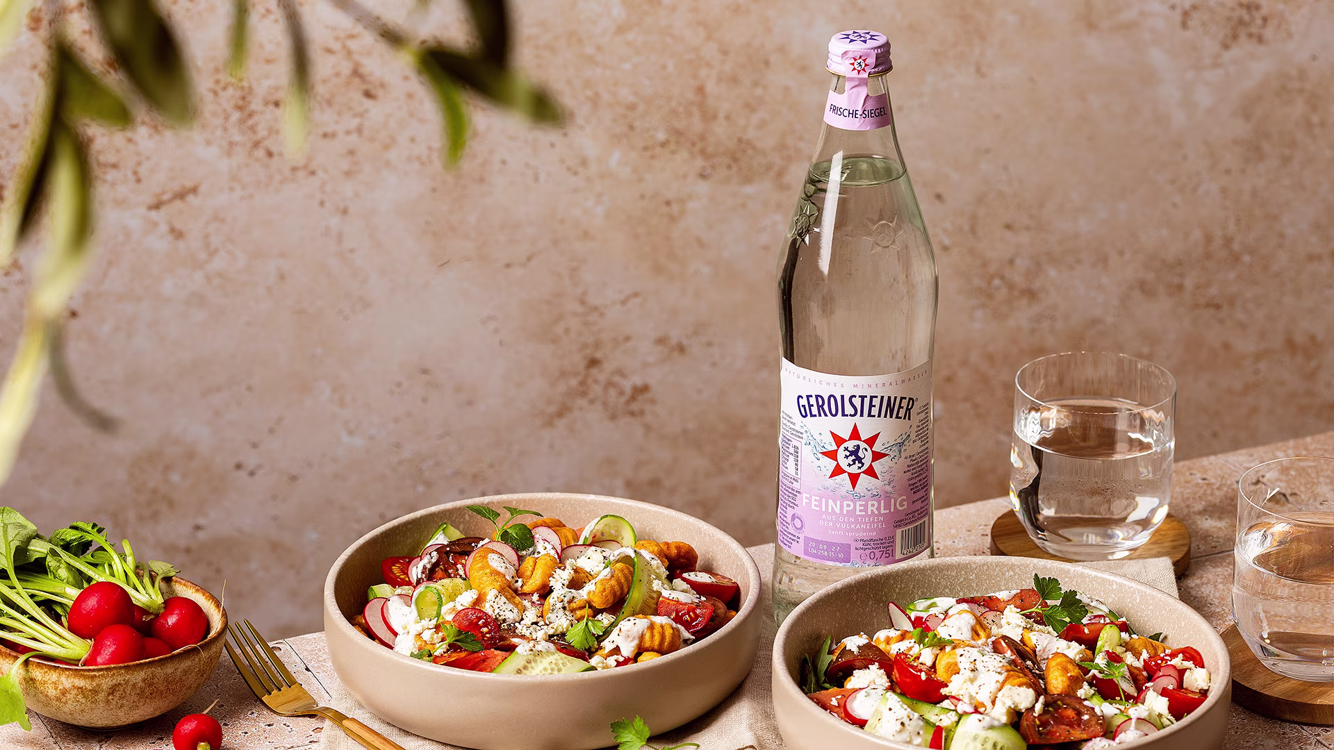 Frischer Salat mit knusprig gebratenen Gnocchi, Radieschen, Tomaten und cremigem Käse in einer sommerlichen Tischszene arrangiert. Dazu eine Flasche Gerolsteiner Feinperlig und Gläser Wasser für einen leichten, mediterranen Genussmoment.