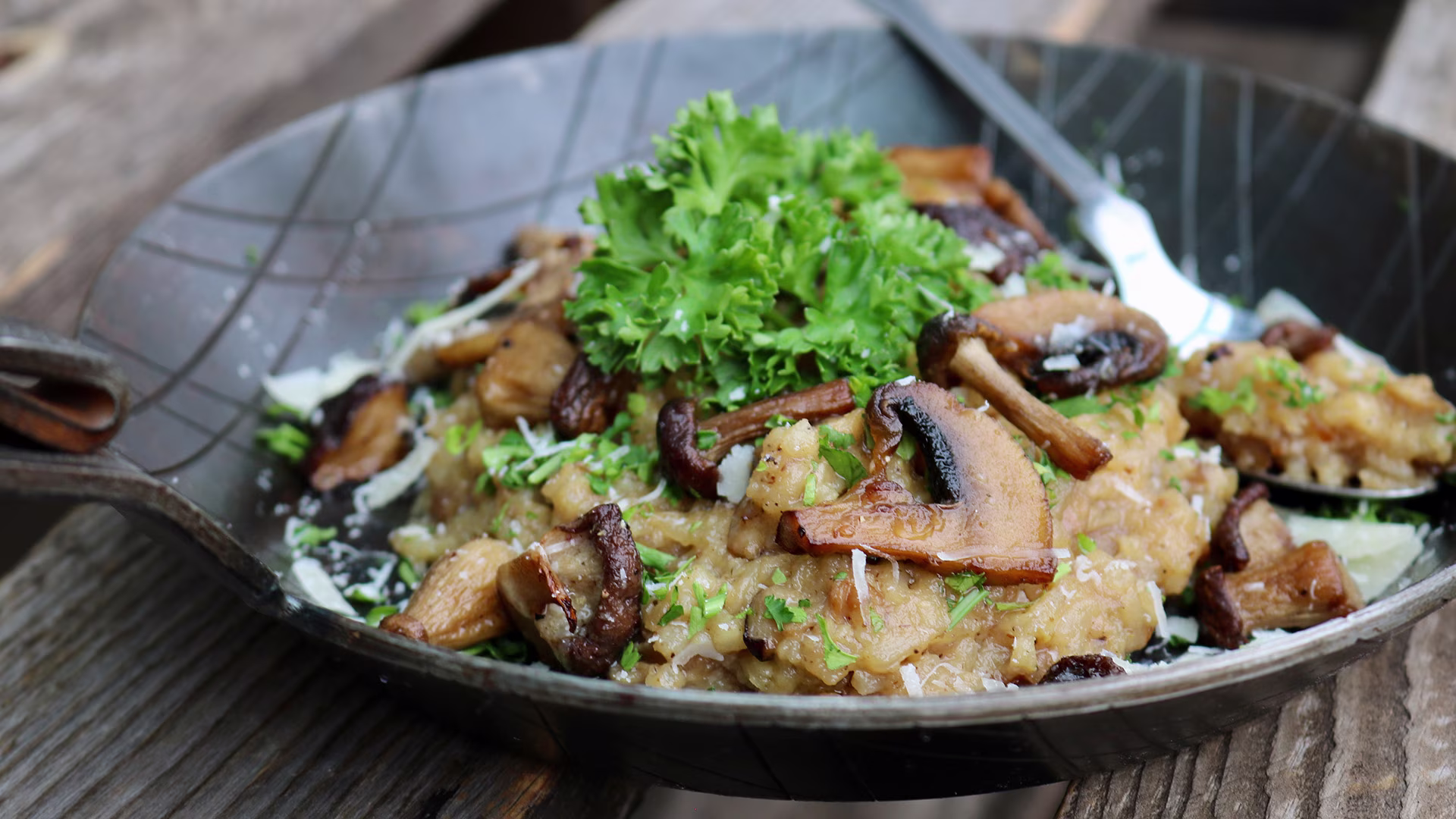 Cremiges Pilz-Risotto mit gebratenen Champignons und Kräuterseitlingen, garniert mit frischer Petersilie und feinen Parmesanspänen, serviert in einer rustikalen schwarzen Pfanne.