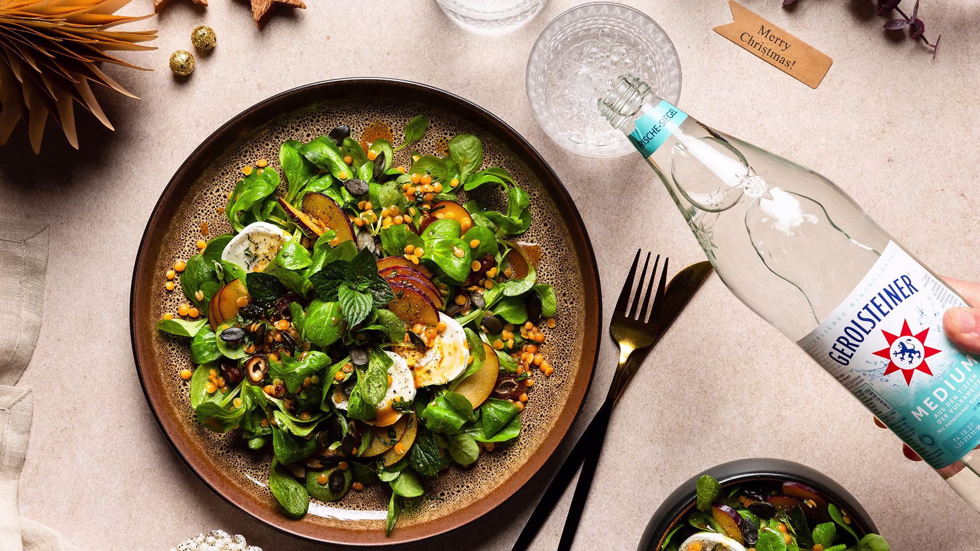 Ein Teller mit Linsen-Feldsalat, Ziegenkäse und Pflaumen liegt festlich angerichtet auf einem Tisch. Eine Hand schenkt Gerolsteiner Medium in ein Glas ein, goldene Deko und ein „Merry Christmas“-Kärtchen sorgen für weihnachtliche Stimmung.
