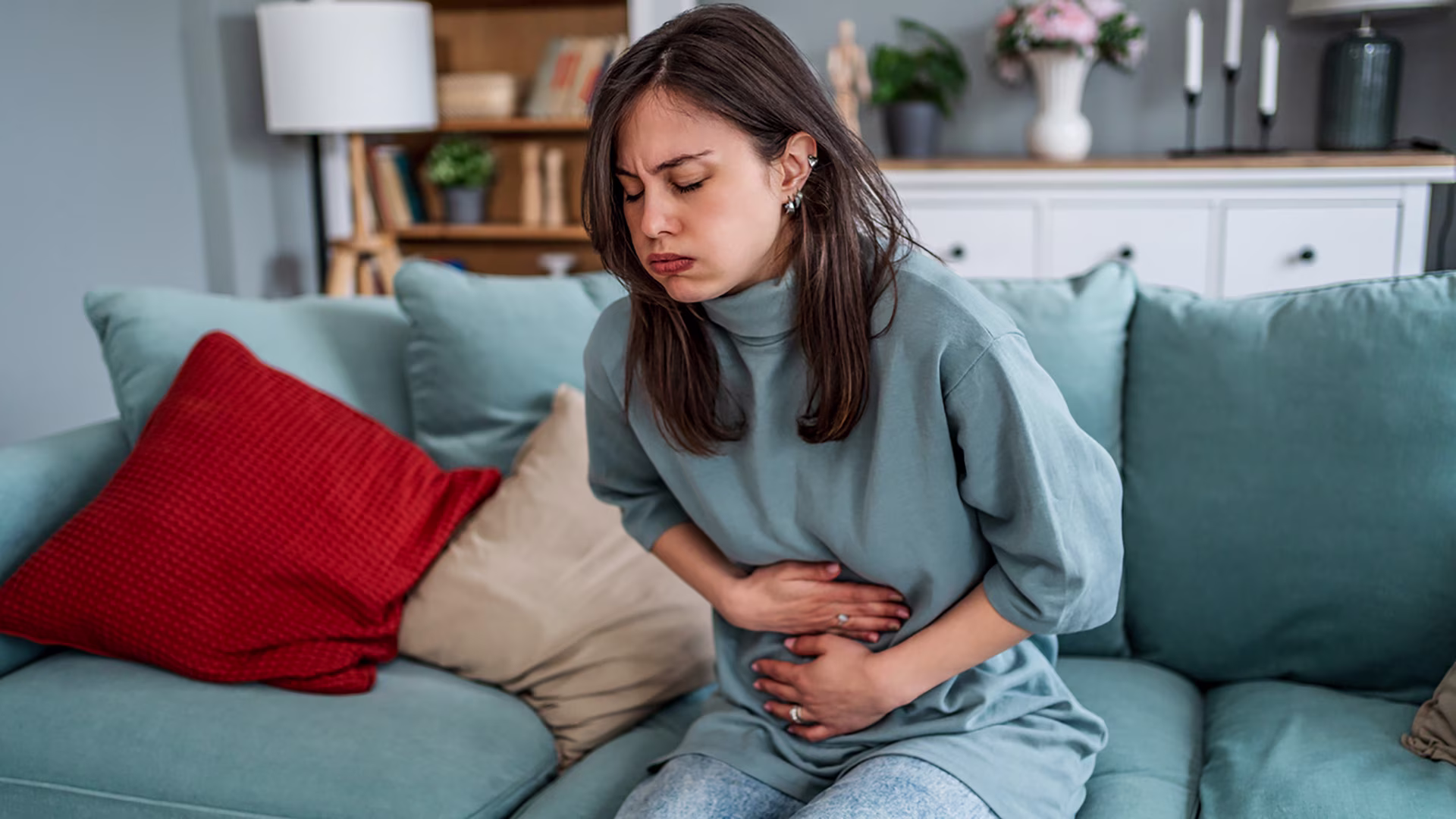 Junge Frau sitzt gekrümmt auf dem Sofa und hält sich den Bauch.