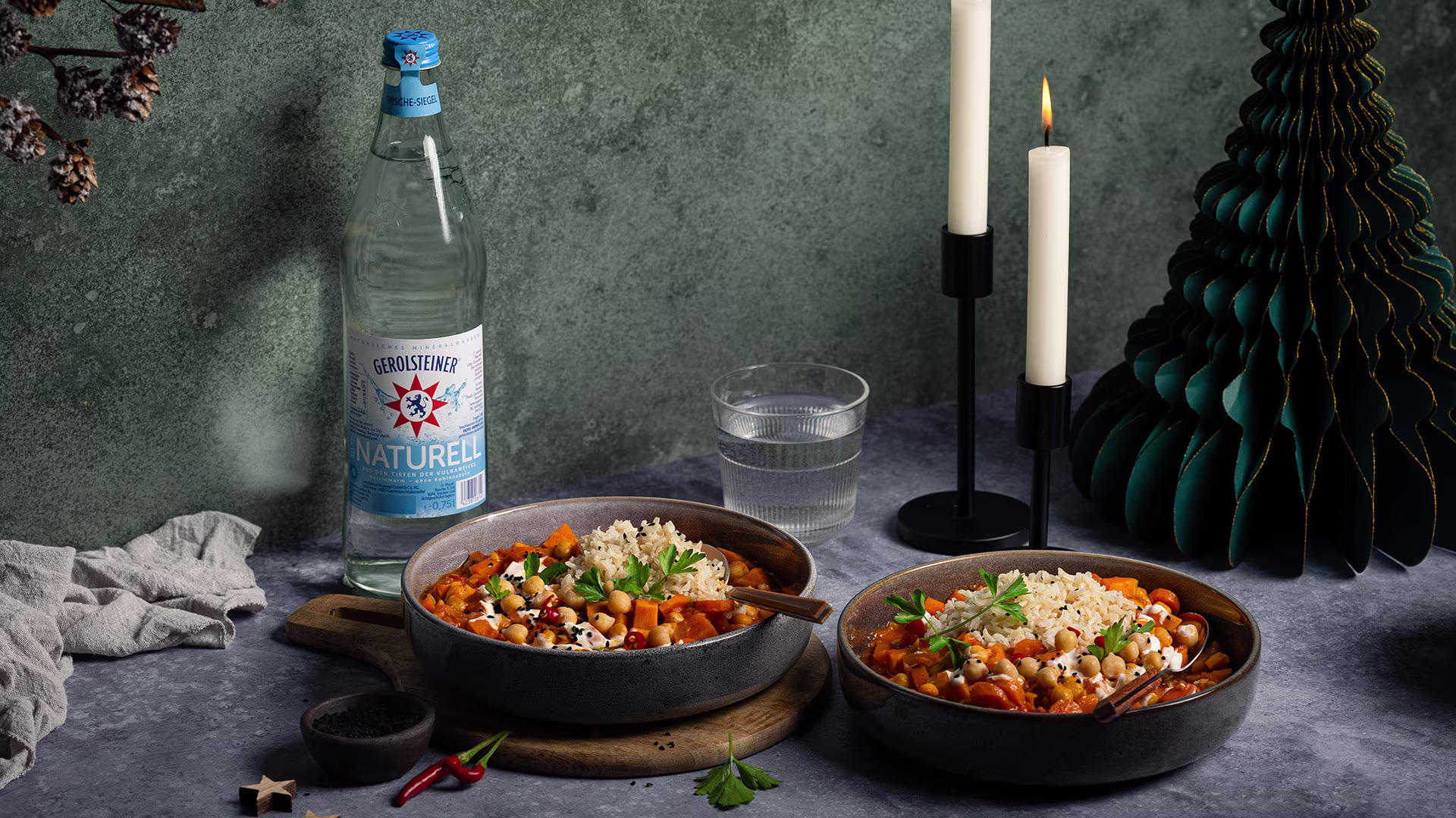 Eine Flasche Gerolsteiner Naturell steht neben zwei Schalen mit Kichererbsen-Eintopf und Reis auf einem gedeckten Tisch. Brennende Kerzen und grüne Dekoration schaffen eine gemütliche, winterliche Atmosphäre.