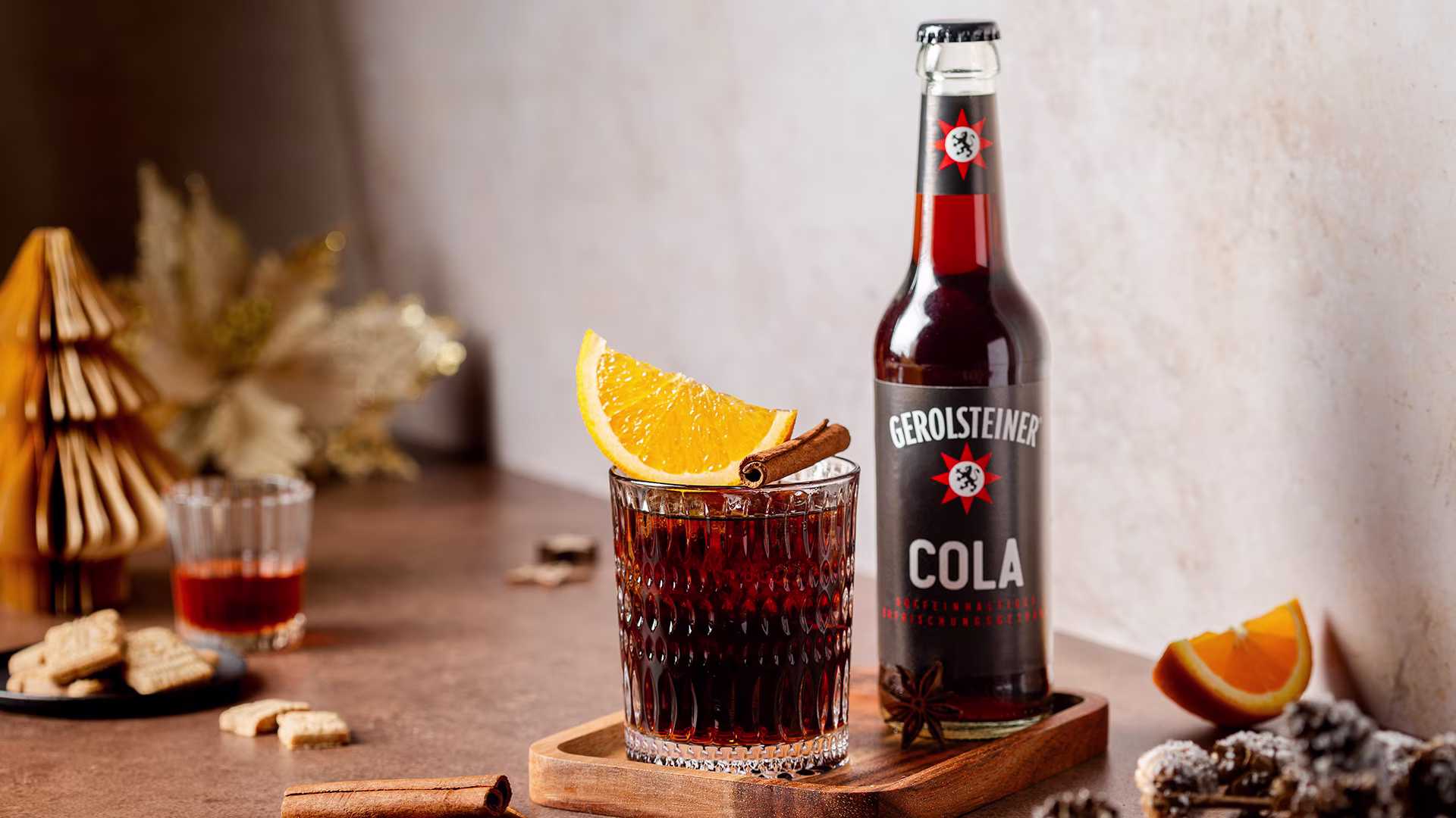 Eine Flasche Gerolsteiner Cola steht neben einem Glas mit Cola, garniert mit einer Orangenscheibe und einer Zimtstange. Auf dem Tisch liegen Gewürze und Weihnachtsgebäck, im Hintergrund sorgen Dekorationen für eine festliche, warme Stimmung.