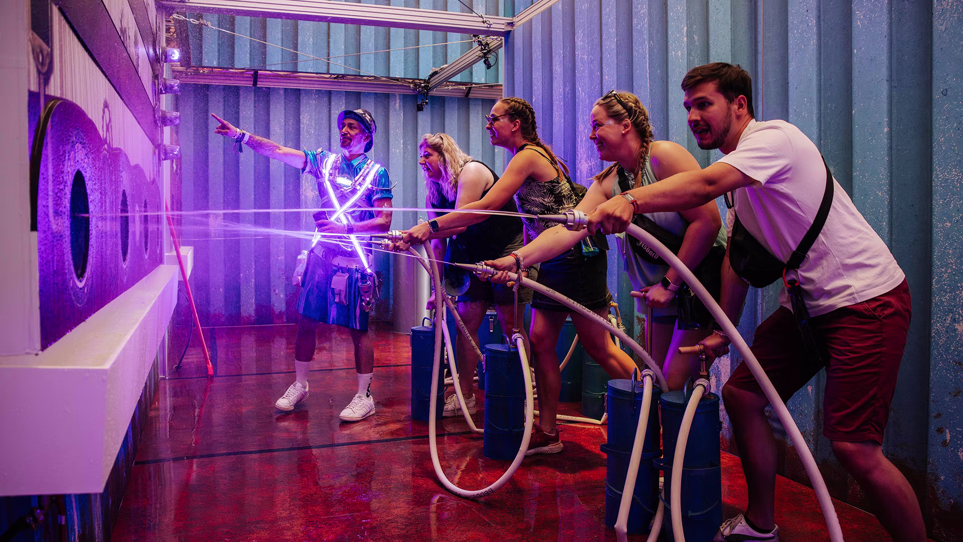 Festivalbesucher zielen mit Wasserschläuchen auf Zielscheiben, angeleitet von einem Promoter im leuchtenden Brandlöscher-Outfit – actionreiche Spielstation in Container-Optik.