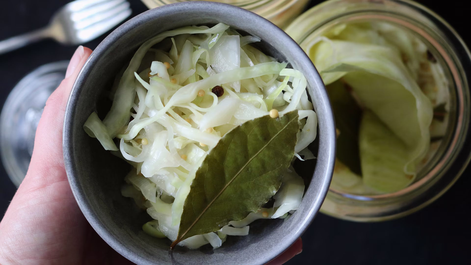 Eine Schüssel mit hausgemachtem Sauerkraut, garniert mit Lorbeerblatt und Senfkörnern. Im Hintergrund sind Einmachgläser mit weiterem Sauerkraut und Besteck zu sehen. Die Szene vermittelt einen frischen, handgemachten und natürlichen Eindruck.
