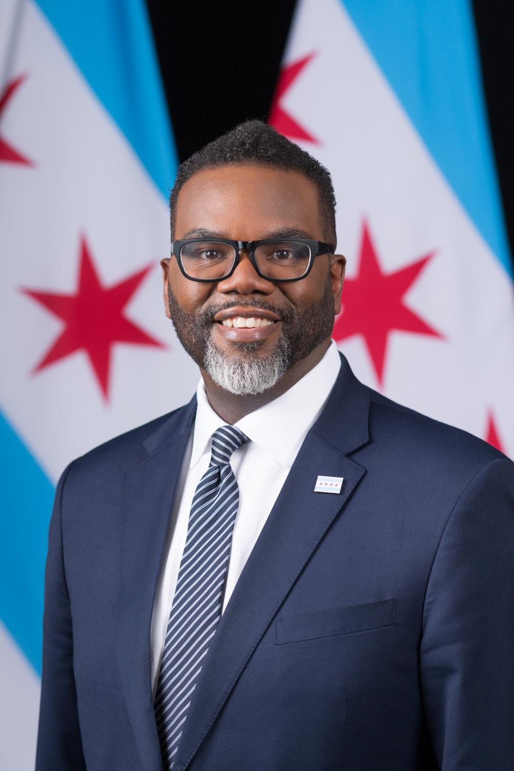 A photographic portrait of a Black man with short, cropped hair and salt and pepper beard smiling for the camera. He wears glasses and a suit and tie. Behind him is part of the Chicago city flag, with 6-pointed red stars and blue stripes.