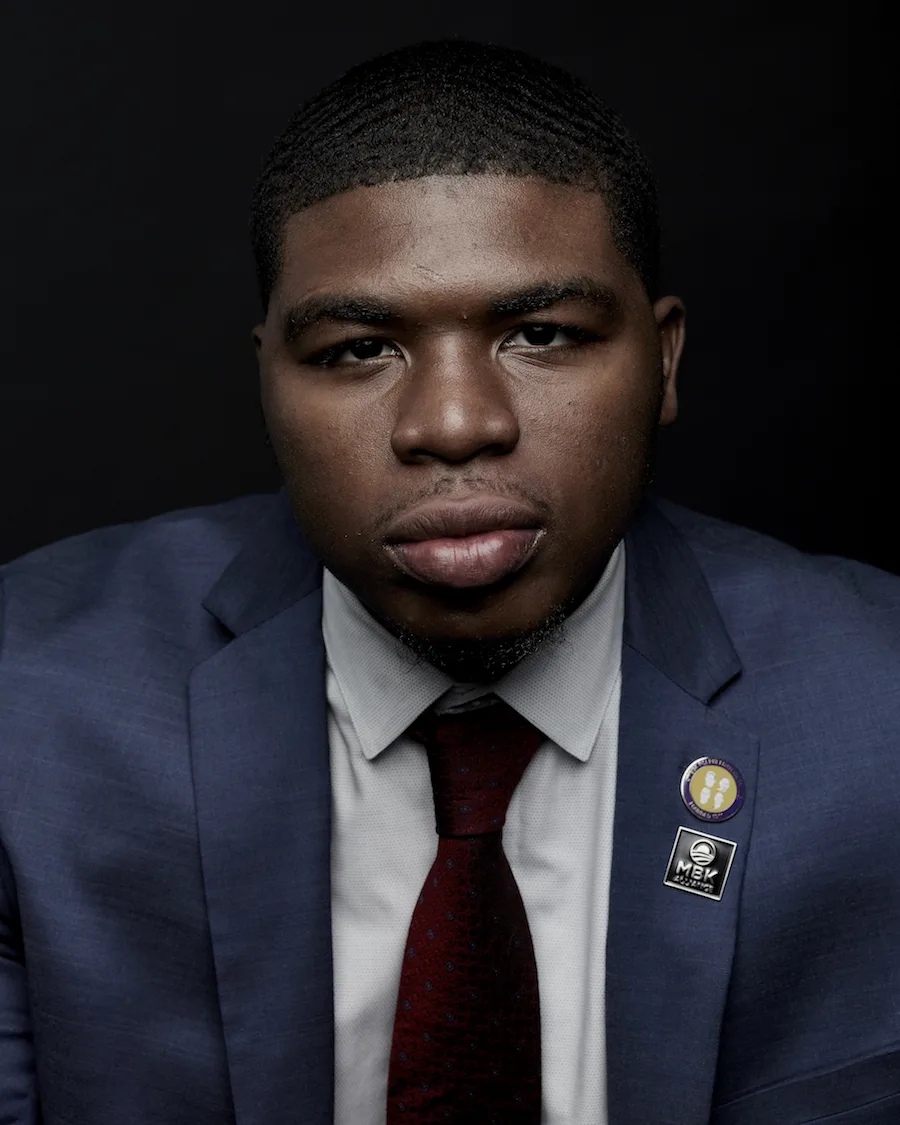 Noah McQueen, a Black man with a deep skin tone, stares into the camera. He is wearing a navy suit with a MBK pin.