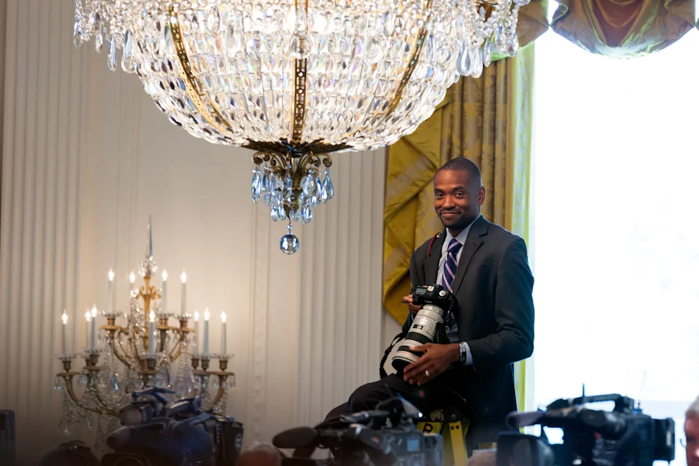 Image of Lawrence Jackson in the White House. Lawrence has a deep skin tone and is wearing a gray suit with a striped tie. He has dark, closely cut hair. He is standing in a room with two large chandeliers with a table in front of him. On the table are several cameras. In his hand is a camera that is hanging around his neck.