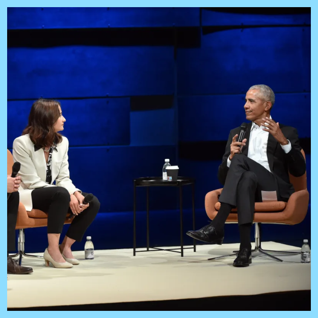 President Obama sits on stage and speaks into a microphone wearing a dark gray suit. Three young people with medium-light skin tones sit to his right looking and listening to him wearing business attire.