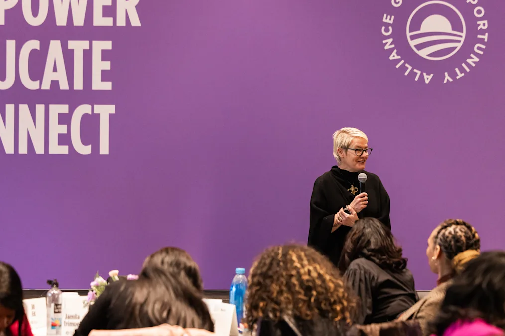 A woman with a light skin tone holds a mic on a stage. In the background, a purple banner reads, “Empower, Educate, Connect.”