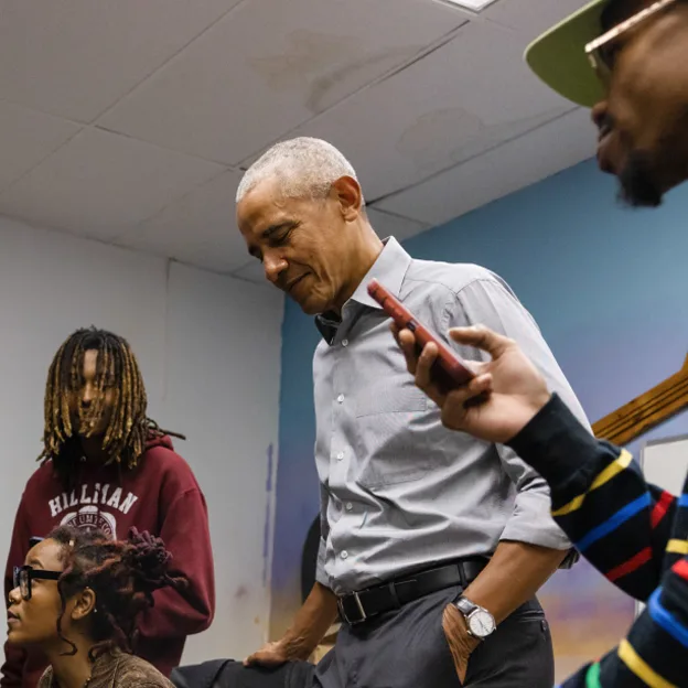 President Obama smiles as he listens to a Black male with a deep skin tone rap from his phone. He is wearing glasses and a neon green cap. Two males with deep skin tones are in the background. 