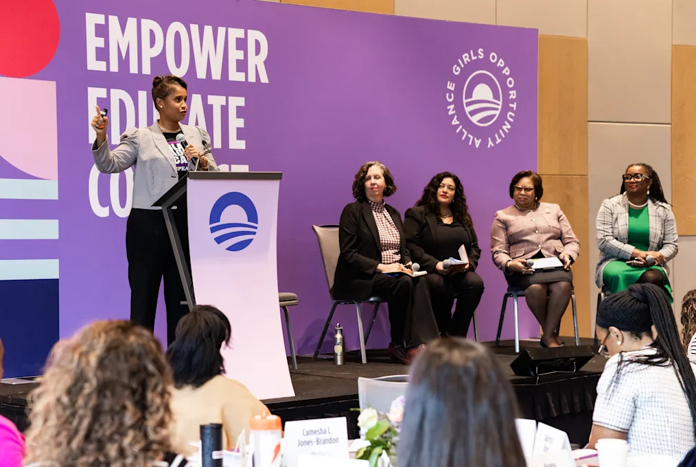 A woman with a medium dark skin tone and short brown hair speaks behind a podium. Four women with a range of light to dark skin tones sit on the stage alongside her. All are dressed professionally. A purple backdrop behind her reads, “Girls Opportunity Alliance” and “Empower, Educate, Collaborate.”