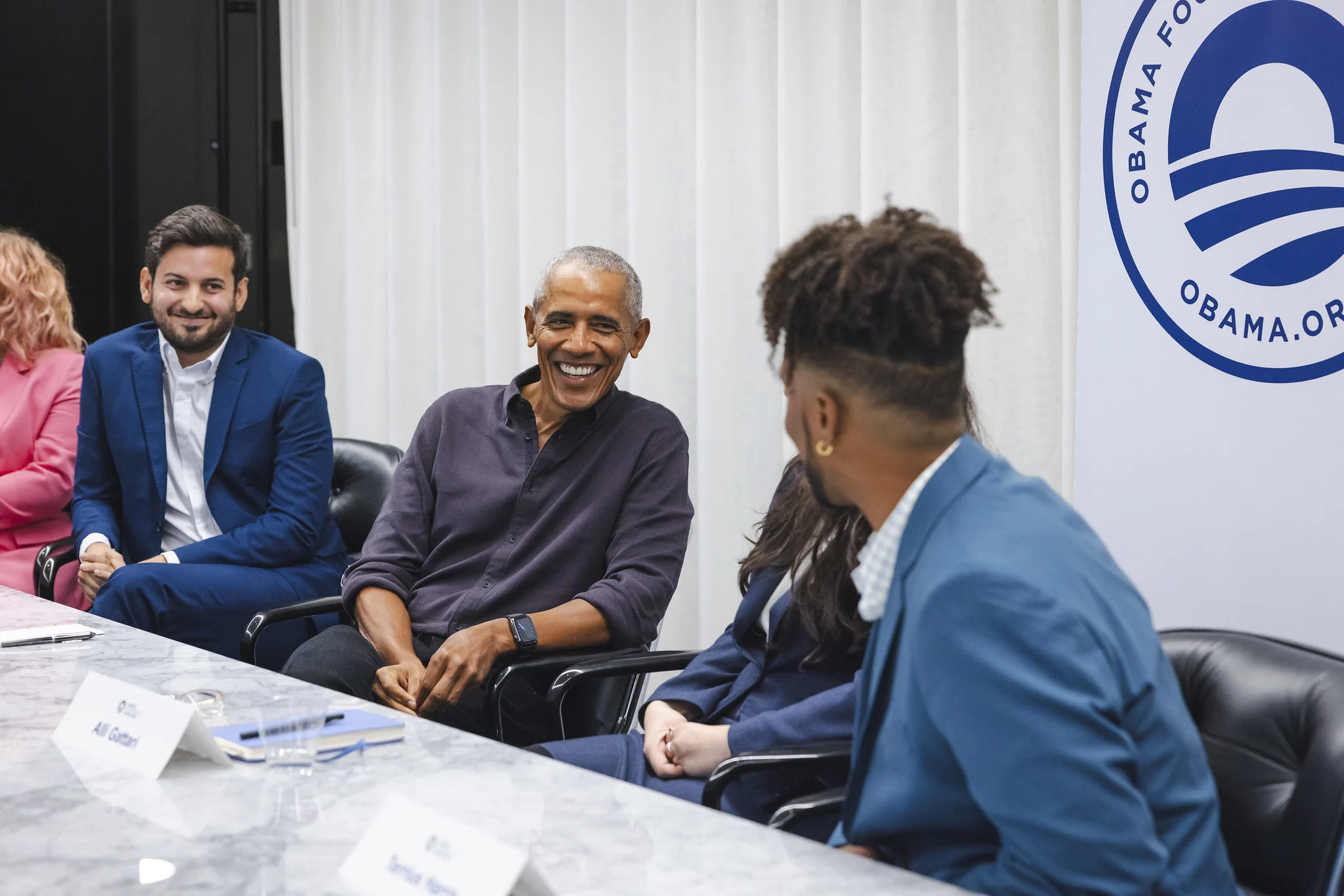 President Obama sits in between a group of people with a range of ages and light to dark skin tones. President Obama is wearing a dark purple button up. In the background is a sign with the Obama Foundation rising sun logo that reads, “Obama Foundation” and “Obama.org.” 