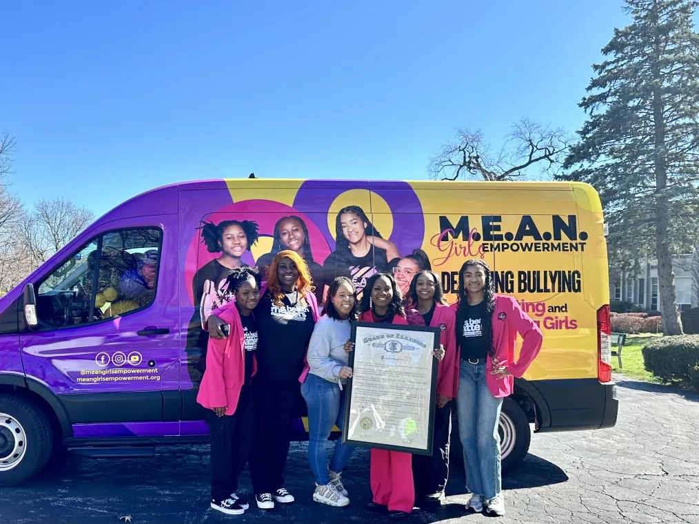 A group of Black women and girls pose with a van wrapped with M.E.A.N. Girls Empowerment branding and images of adolescent Black girls. The group is holding a copy of Governor Prizker’s state proclamation recognizing Girls Health Matters Day. 