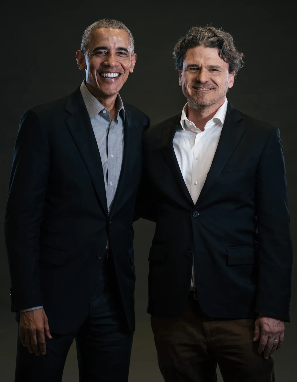 In this picture, President Obama wearing a black suit and gray shirt poses beside
a man with a light skin tone wearing a black suit and a white shirt, while smiling
 at the camera.