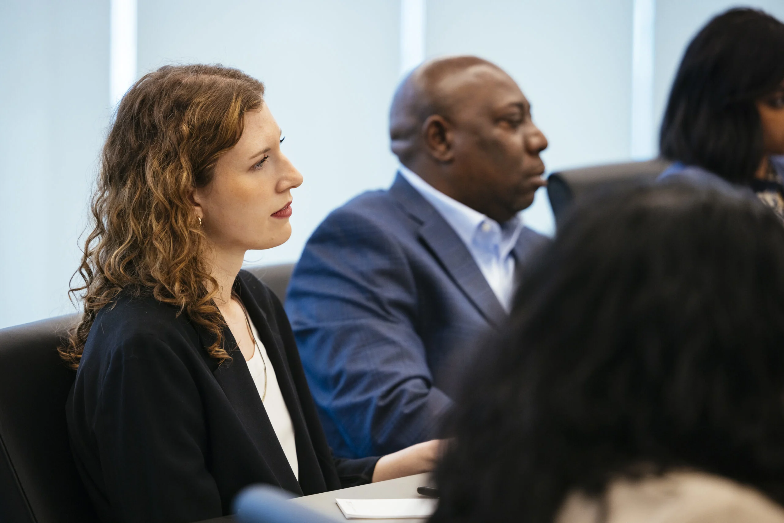 A woman with a light skin tone wearing red lipstick and a black blazer sits at a table next to a man with a medium-deep skin tone in a dark blue blazer.