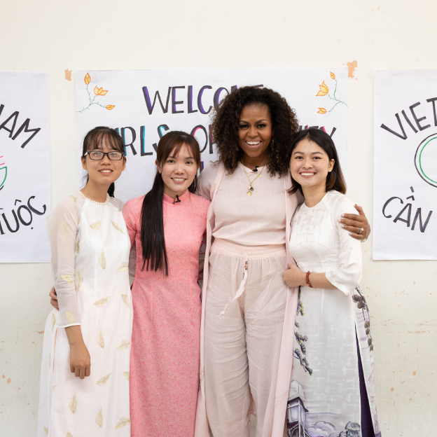 Michelle Obama stands in the center of a photo posing with three other women. Behind the group are three posters. The first woman has an olive skin complexion, is wearing a white dress, glasses, has dark hair and is smiling at the camera. The second woman has olive skin complexion and long dark hair. She is smiling and wearing a long pink dress. The third woman in the picture is Michelle Obama. She has medium deep skin tone, curly hair, and is wearing beige pants, a beige shirt, and beige pants. The fourth woman in the picture has a olive skin complexion, dark hair cut in a bob, and is wearing a white long shirt with flowers on the bottom. 