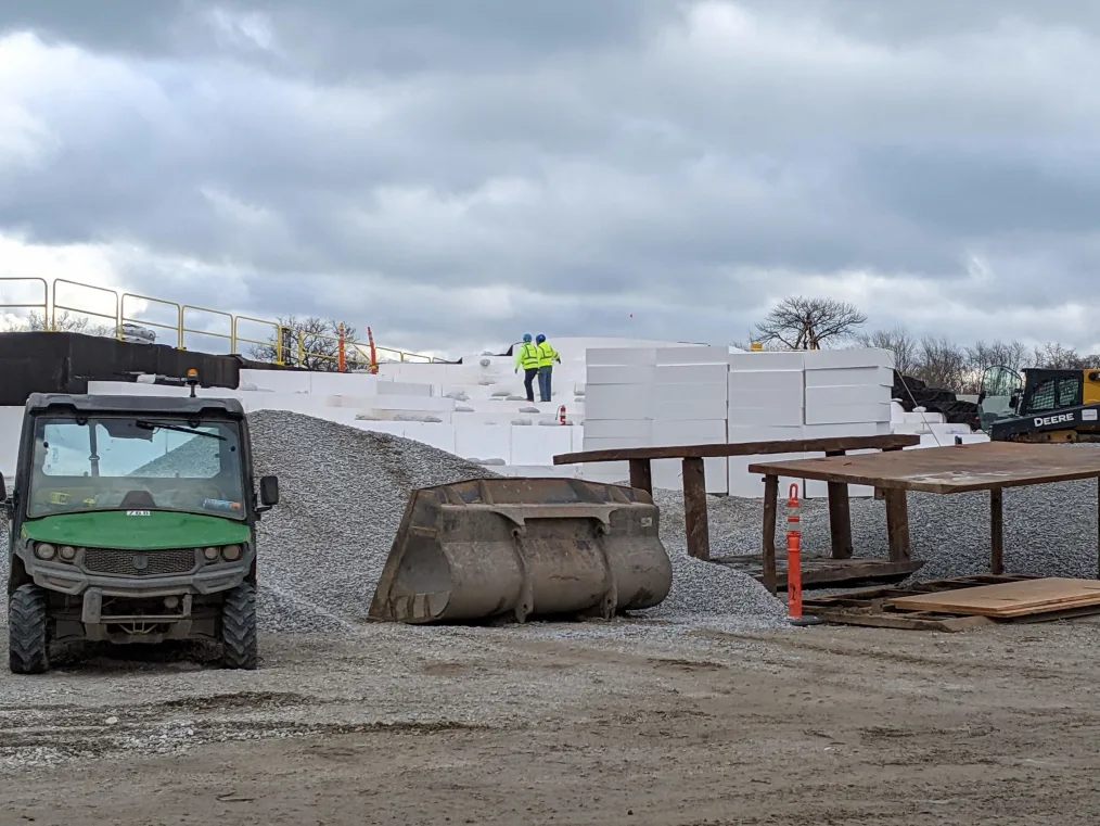 Workers wearing bright safety vests walk and blue hard hats as they walk on large white blocks of geofoam that will form the base of the playground and great lawn. In the foreground, large metal construction implements are strewn about and a green and white golf cart sits to one side.