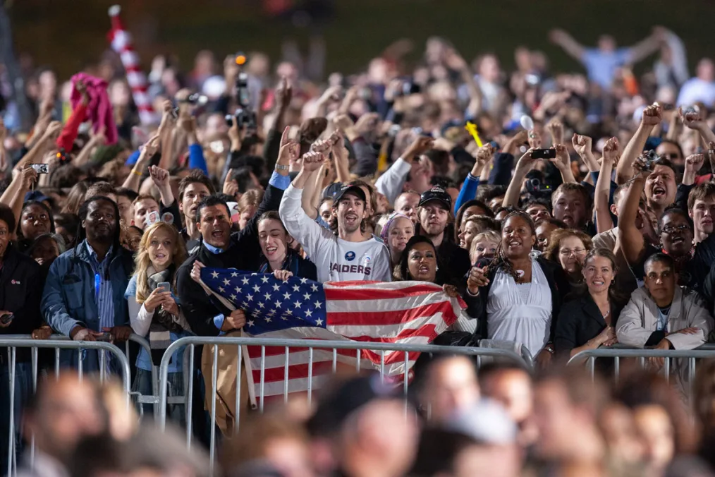 A crowd of people cheering with their arms in the air. At the center is a trio holding an American flag. Other people hold up cameras.