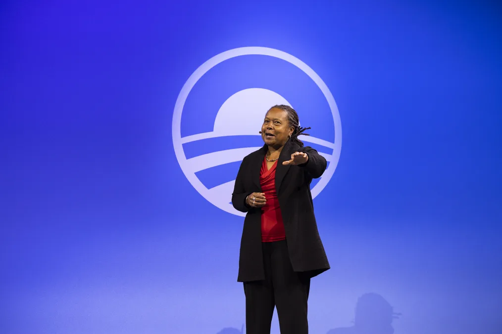 A older woman with medium deep skin stands giving a speech. She has grey and black long locks, a round face, a black jacket, with a red dress shirt underneath and black pants. She has a mic coming from her right ear. She is making hand gestures towards the crowd. In the background is a light blue and dark blue gradient with the Obama Foundation logo in the center.