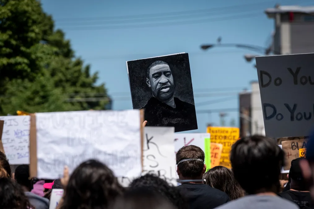 A photo of George Floyd is raised during a protest. A group of people gather in the street with signs. Their backs are to the camera.