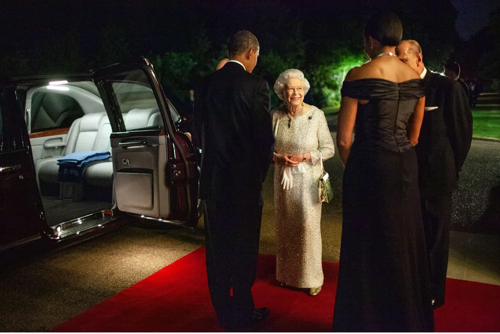 At nightfall, President and Mrs. Obama face Queen Elizabeth II as she smiles at them, sending them off into their car that has its door open. Prince Philip is standing next to the Queen, but only a portion of his profile can be seen.
