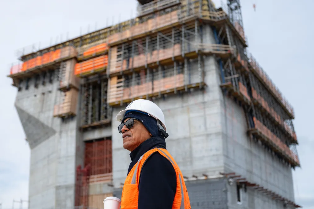 President Obama wears sunglasses, an orange safety vest, white hard hat and holds a cup of tea as he stands in front of a structure at the site of the Obama Presidential Center. A cloudy sky is in the background.