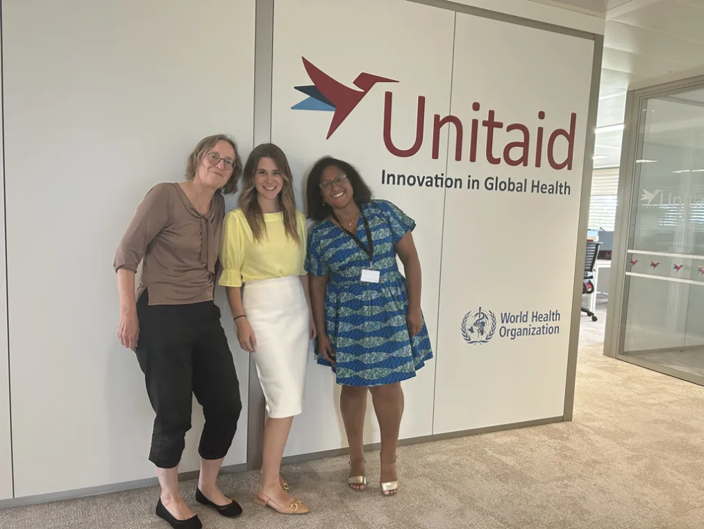 Liza Zaruba, a woman with a light skin tone and long blonde hair, stands in between her supervisors, Karin Timmermans, a woman with a light skin tone and short blonde hair, and Cherise Scott, a woman with a medium dark skin tone and short curly hair. Both are wearing glasses. A sign behind them reads “Unitaid” and “Innovation in Global Health.” Liza is wearing a yellow blouse and white skirt.