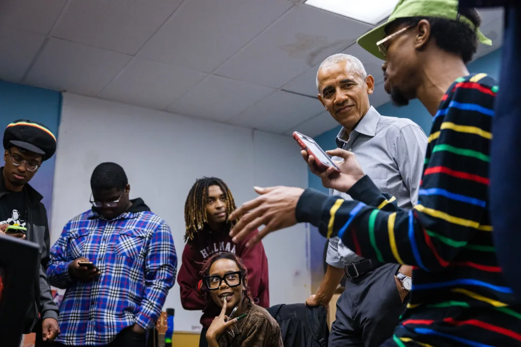  President Obama smiles as he listens to a Black male with a deep skin tone rap from his phone. He is wearing glasses and a neon green cap. Three males and a woman with deep skin tones are in the background. Her finger is on her lip as she listens in. 