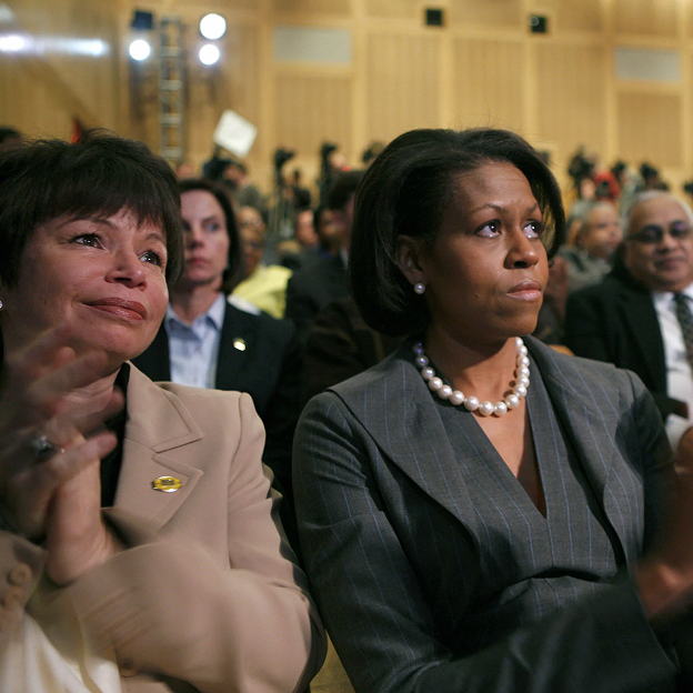 Michelle Obama, her eyes watering with tears and her mouth pursed, sits in a room full of people. Bright spotlights are on the back wall. Everyone looks dressed in formal suit attire.